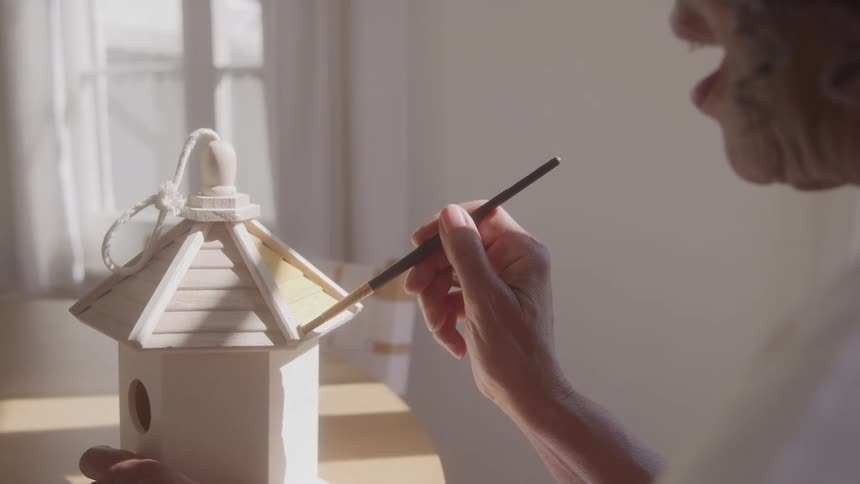 A woman with light colored hair and a white shirt is holding a small unpainted wooden birdhouse and using a brush to paint the roof a light color