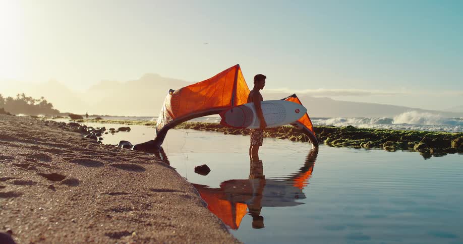 Surfer standing by the sea holds a board and orange sail reflected in the water
