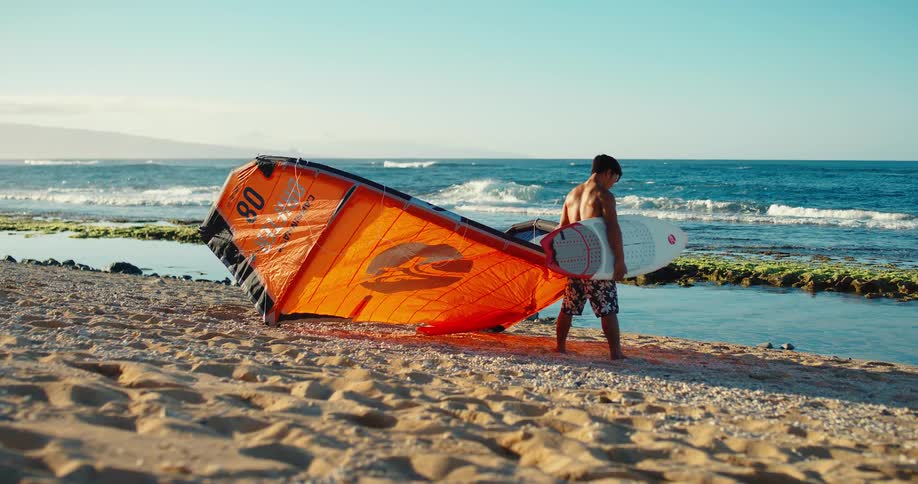 Man on the sand holds a surfboard and a kitesurf kite while looking at the sea