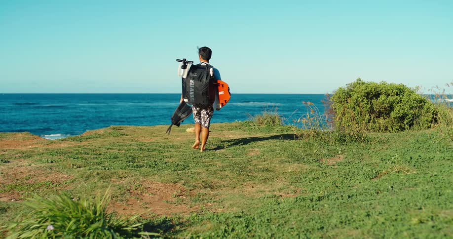 Man walks across the grass toward the sea carrying a water sports backpack