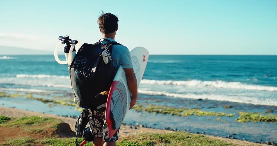 Surfer looks at the sea holding a surfboard with a backpack on his back