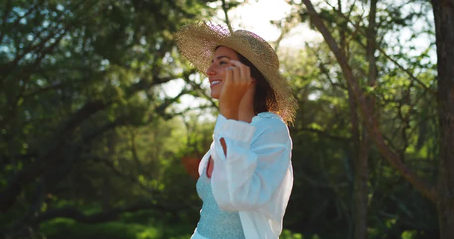 A woman wearing a large straw sun hat a light blue top and a white button down shirt is standing in a forest clearing with her eyes closed smiling as she looks up toward the sun