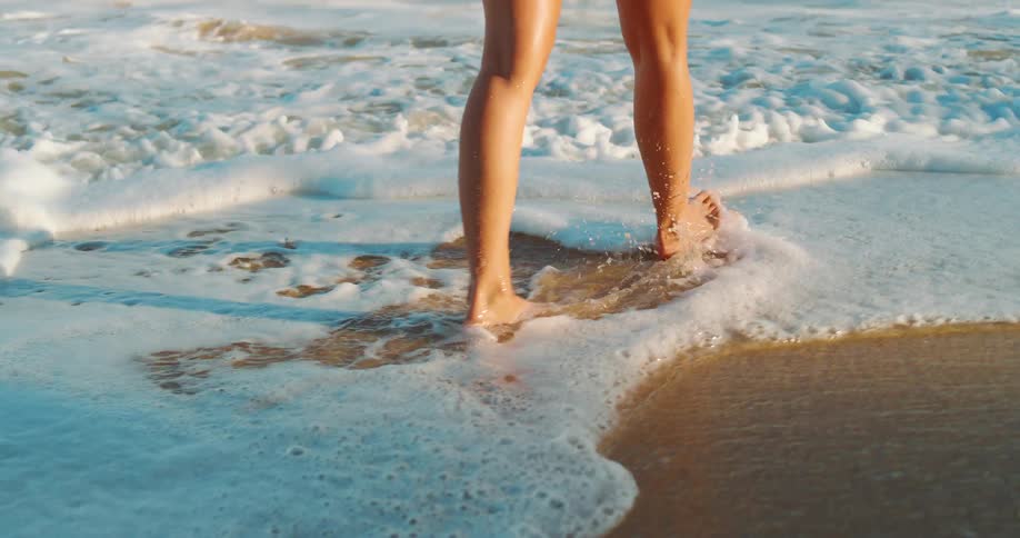 A woman slowly walks along a beach a ocean water washes over her feet