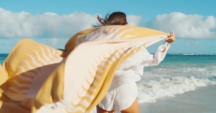 A woman walks along a beach on a windy day holding a beach blanket behind herself
