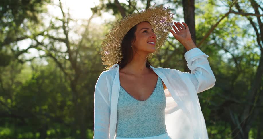 A woman wearing a large straw sun hat a light blue patterned top and a white button down shirt is standing in a sunlit forest clearing with her hands touching her hat as she looks to the side with a calm expression