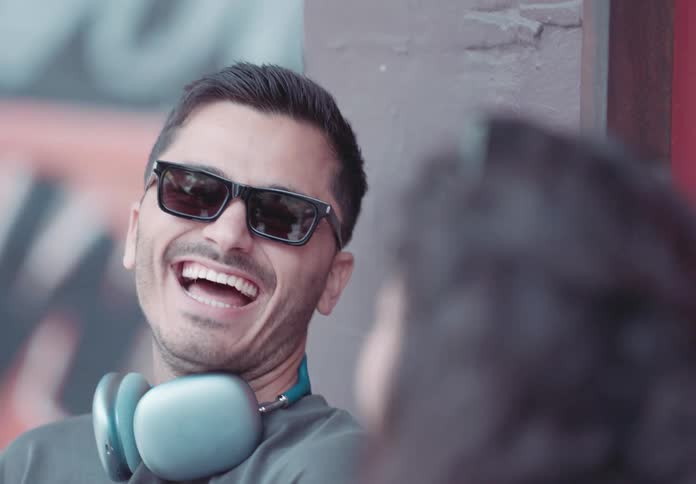 A man with a wide smile and a pair of light blue wireless headphones around his neck is talking at a person with his dark glasses reflecting the surroundings