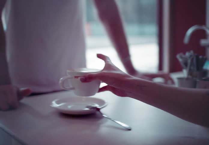 A young man wearing a white tshirt standing inside a caf is served a cup of coffee with a spoon on the side by the saleswoman at midday