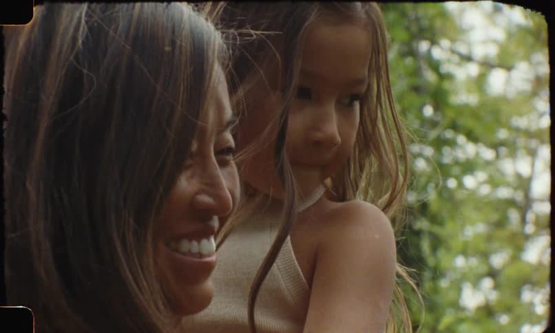Smiling woman holds a child in her arms while both look forward in an outdoor setting with trees in the background
