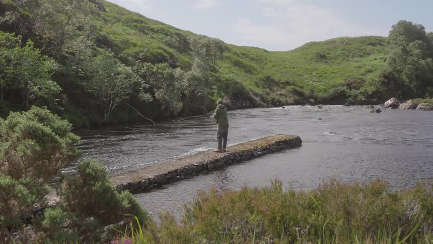 A man in a jacket stands on a narrow stone pier casting a fishing line into a river surrounded by lush green hills