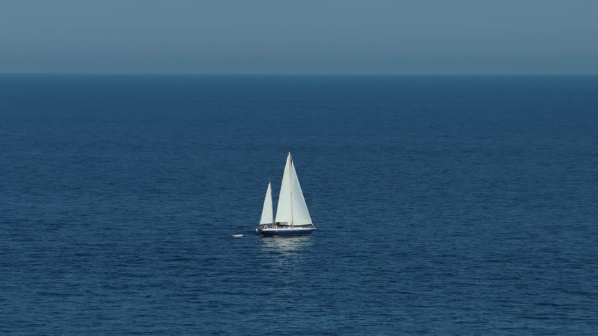 A lone sailboat with white sails moves slowly across the vast deep blue open ocean under a hazy sky