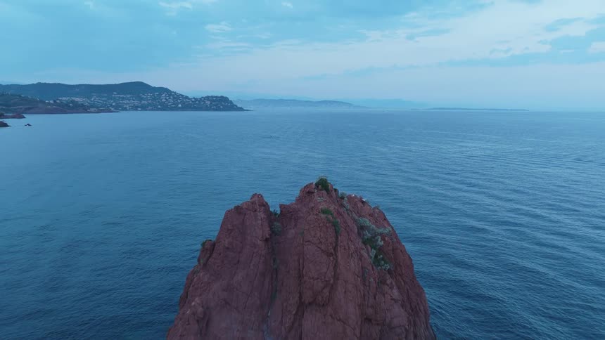 An aerial shot flies forward over a large reddish rock formation protruding from the sea with a coastal city visible in the background at dusk