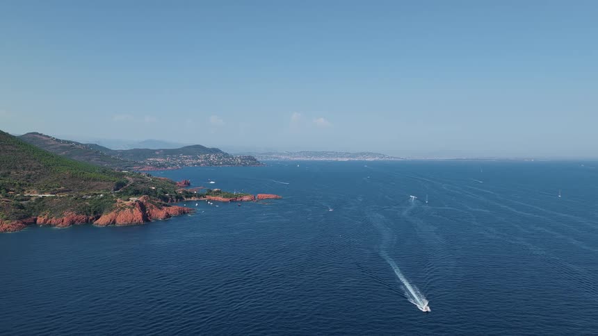 A wide static aerial shot captures a speedboat cruising across the ocean alongside a scenic mountainous coastline dotted with towns