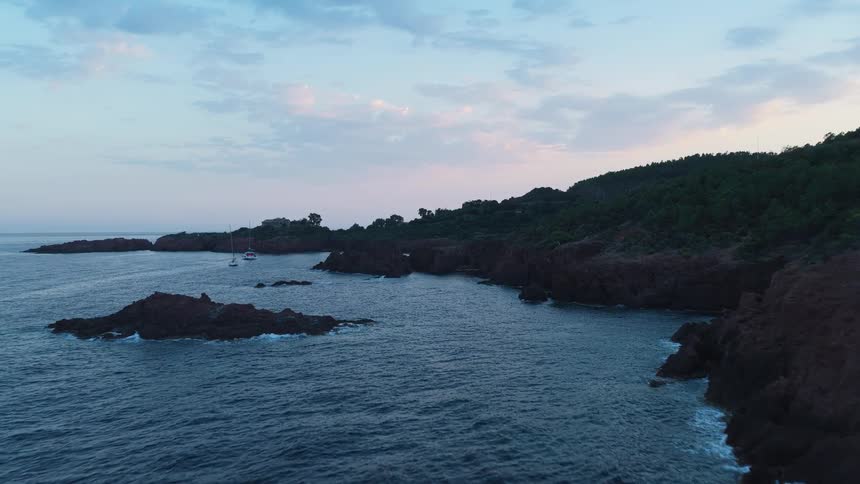 A sweeping aerial shot flies along a rugged jagged coastline with reddish cliffs and forested hills at dusk revealing sailboats anchored in a cove