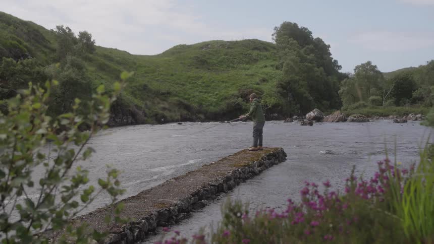 A man stands at a river and uses a long fishing pole to fish