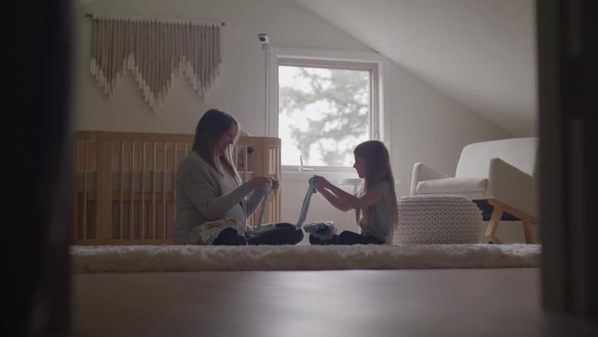 A mother and daughter are sitting on a light colored rug in a babys nursery smiling and talking as they hold up and look at small blue and white articles of baby clothing