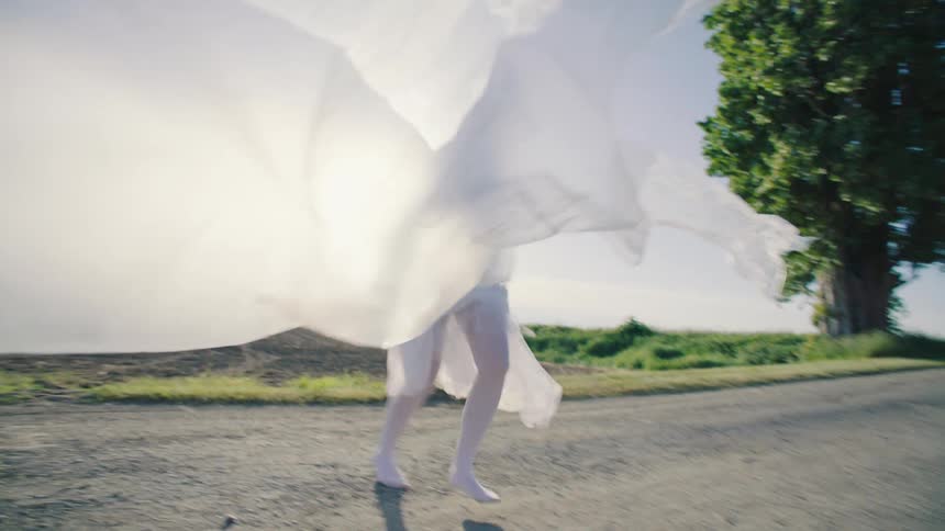 A bride wearing a white gown with a large attachment flowing in midair from the wind as she walks across the road to a grass field with two tall trees at midday