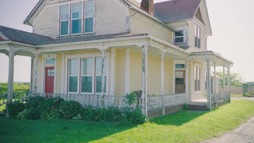 A shot pulls back to reveal a large old yellow farmhouse with a weathered wraparound porch on a bright sunny day