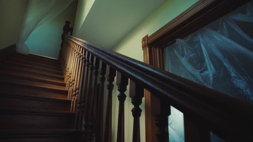 A pointofview shot moves slowly up a dark wooden staircase inside a house that is being renovated with plastic sheeting covering a nearby doorway