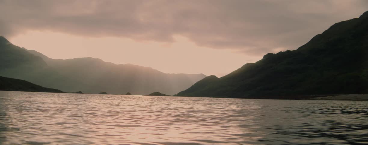 A calm body of water with small ripples on its surface is surrounded by a pair of large dark mountains under a cloudy light sky