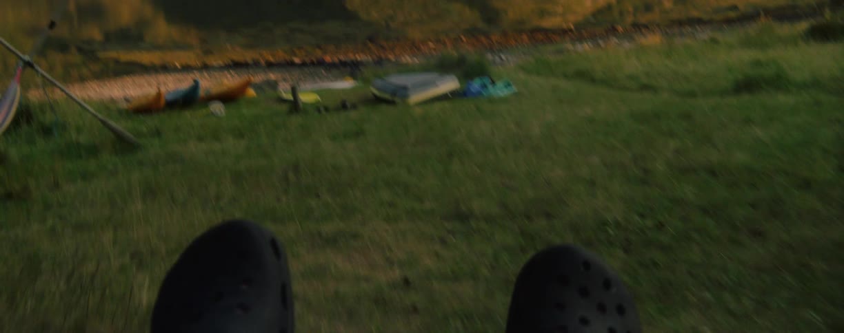 A view from a persons perspective shows the tips of their black shoes in the foreground as they sit on a swing in a green grassy field with a calm lake and a large grassy hill visible in the distance