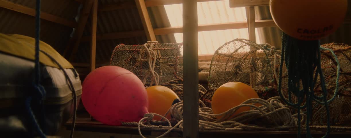 A variety of fishing equipment buoys coiled ropes and woven crab pots all illuminated by bright light shining through a window behind them inside a shed
