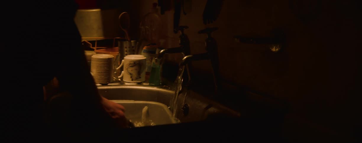 A view of a kitchen sink in a dark room shows a persons hand washing a plate while a water droplet drips from a faucet into the basin