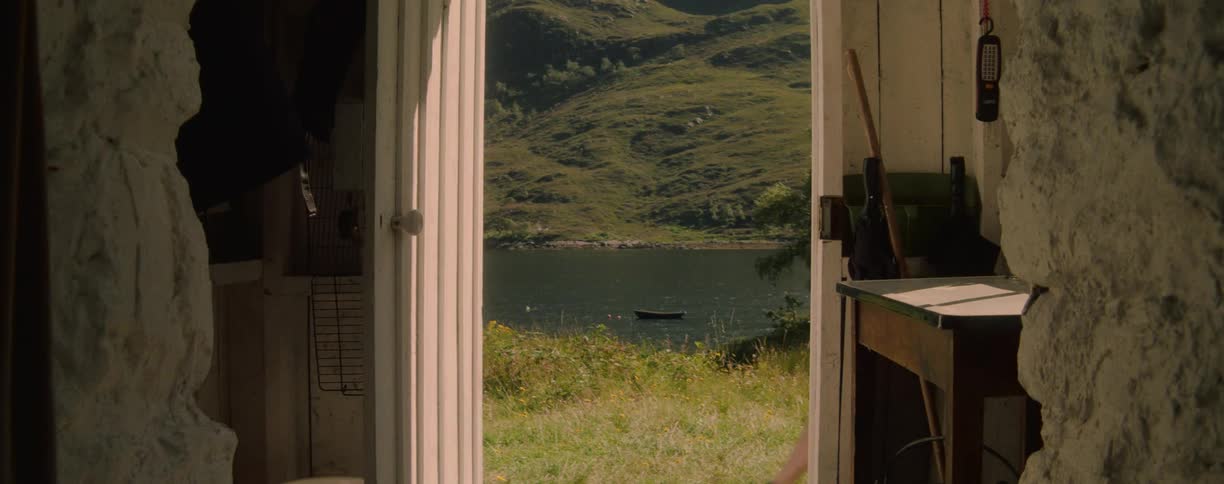 A person wearing a dark shirt and tan pants is opening a light colored wooden door to a cabin and stepping out into a sunlit green landscape with a lake and large hills in the background