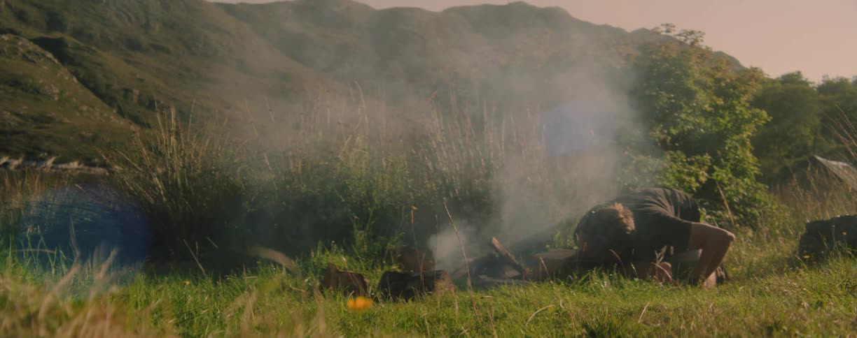 A person tends a small campfire in a grassy mountain landscape