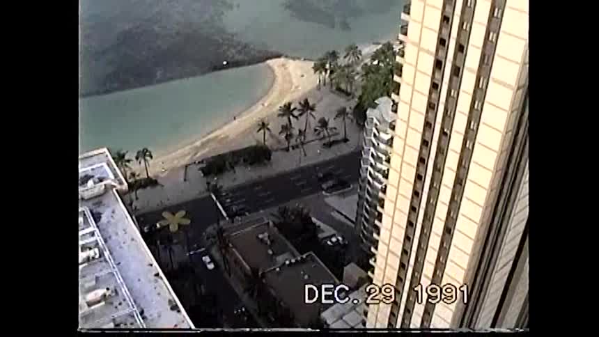 A grainy video from an elevated position shows a wide sandy beach with people and palm trees with the camera panning down a tall building and then out to the horizon to reveal the ocean with a single sailboat with a digital timestamp