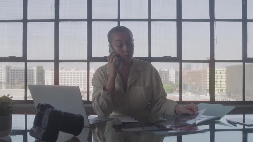 A woman with very short dark hair wearing a neutral colored collared shirt is sitting at a large glass desk in a sunlit office holding a phone to her ear while she looks at a document in her other hand