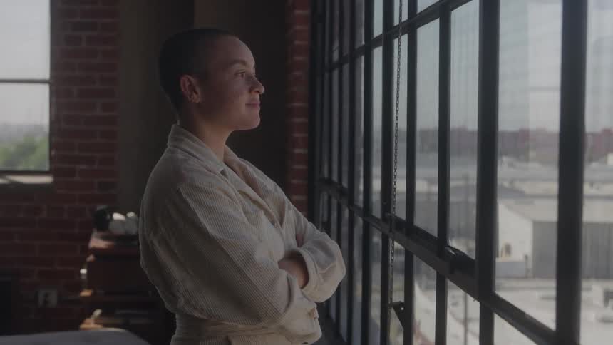 A woman in a robe gazes out a loft window arms crossed