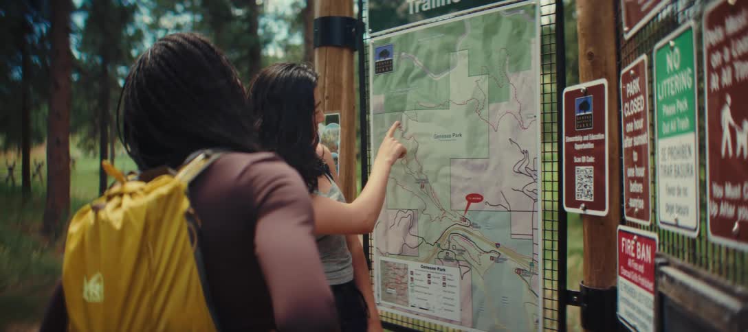 Two young women with dark hair are standing in front of a trail map with one woman pointing to a spot on the map while the other wearing a yellow backpack listens intently