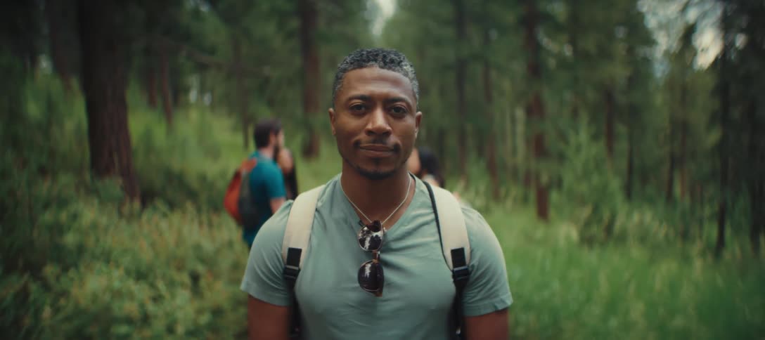 Man with a backpack and sunglasses hanging on his shirt is on a forest trail