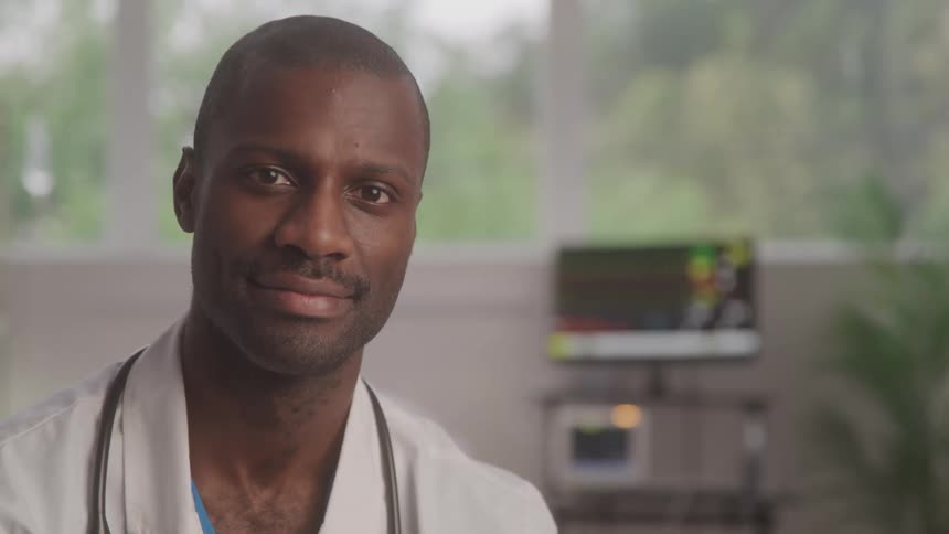 A man with a shaved head and a faint beard wearing a white lab coat over a light blue scrub top is looking to the left and then turning to face the camera with a subtle smile
