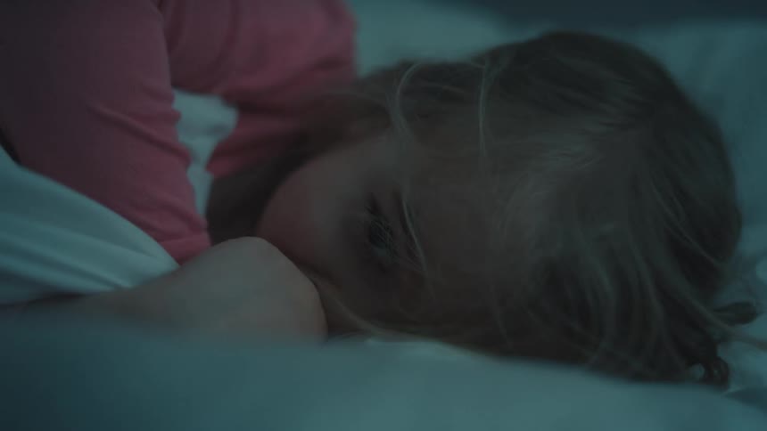 The video shows a child sleeping peacefully in bed the child is wearing a pink shirt and is mostly covered by a white blanket with colorful tassels the childs eyes open for a moment and they briefly stir before drifting back to sleep