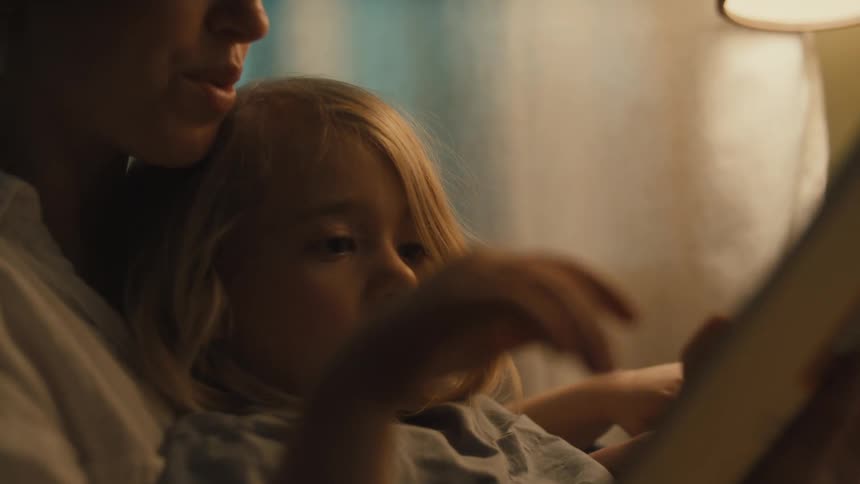 A closeup of a woman and a child sitting together as the woman reads from a book and the child intently listens