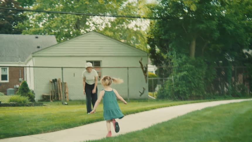 A young girl with blonde pigtails runs through a grassy yard towards a woman while a boy rides a bicycle past them on a paved path
