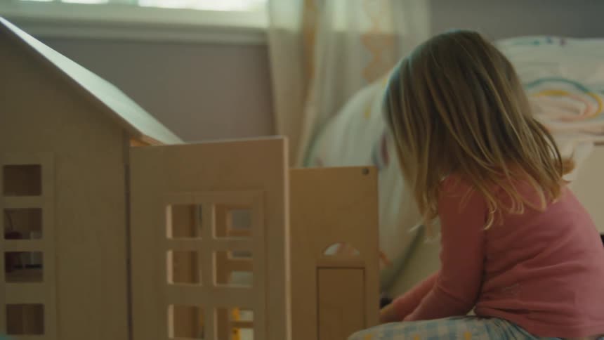 A young girl with blonde hair sitting on the floor intently playing with a wooden dollhouse in her bedroom