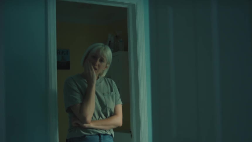 A woman with almost white hair walks towards a room looks inside brings her hand to her mouth in sadness then leaves
