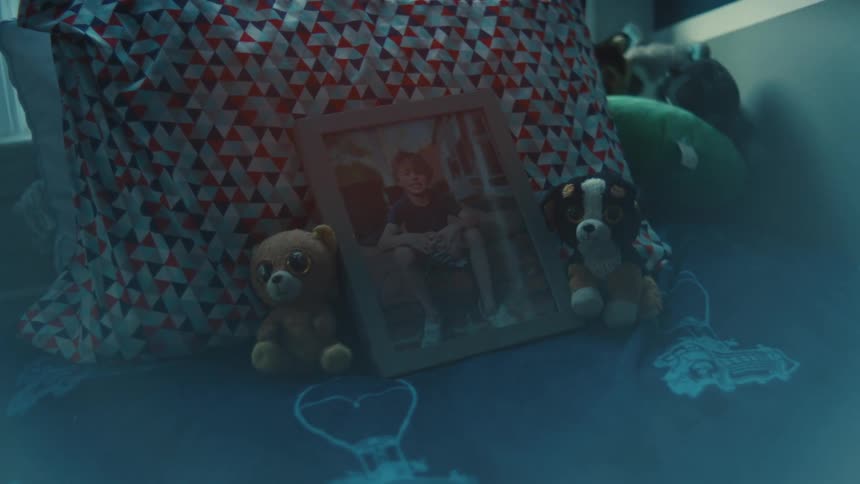 A closeup of a childs bed in a dimly lit room where a framed photograph stands propped up against a patterned pillow surrounded by a couple of stuffed animals