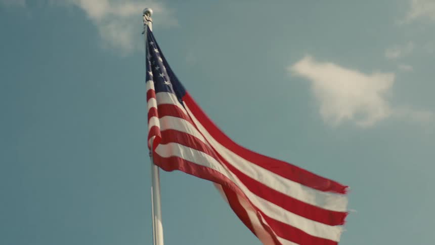 A closeup of a large american flag waving majestically from a white flagpole against a bright blue sky with scattered white clouds