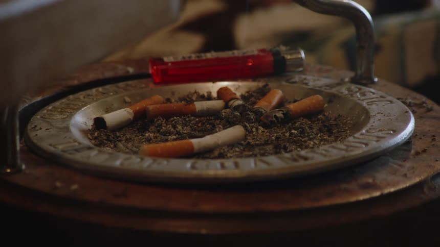 A round metal ashtray filled with dirty cigarette butts and ash sitting on a wooden table beside a red lighter with a metallic top