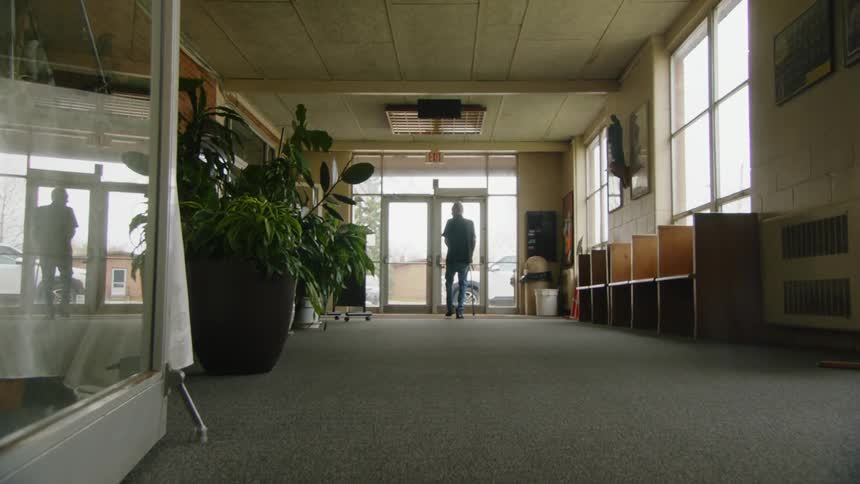 The video shows an elderly man walking away from the camera in a long hallway he is using a cane to walk and is headed toward a set of glass doors at the end of the hall he reaches the doors opens one and exits the building