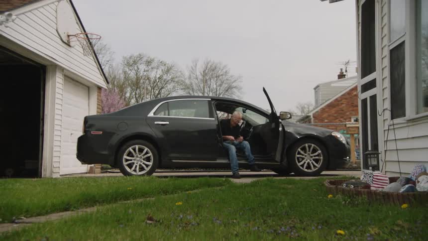 Man sitting at the open door of a black car holding something in his hands