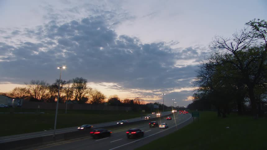 From a static vantage point on a grassy hillside cars and semitrucks with their lights on drive along a busy highway at dusk under a dramatic cloudy sky