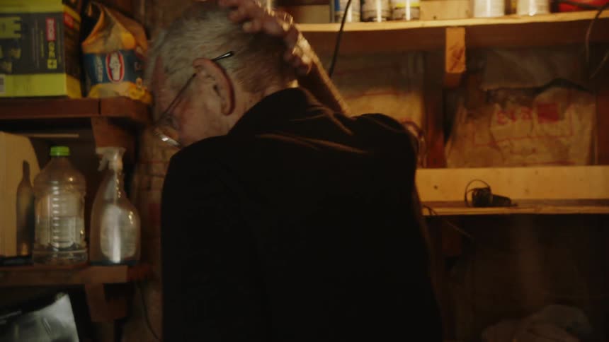 A view from behind shows an elderly man with short white hair and eyeglasses walking through a dark cluttered garage or shed with a single light bulb hanging from the ceiling and shelves filled with miscellaneous items in the background