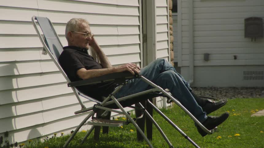 A man with gray hair and glasses sits in a gray folding chair on a green lawn beside a white house smoking a cigarette before taking it out of his mouth holding it in his right hand and exhaling a cloud of smoke