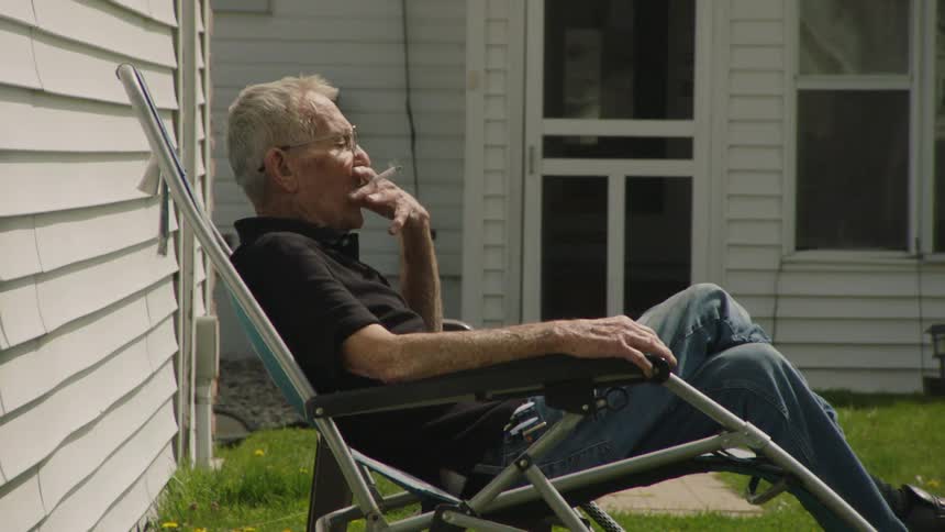 A sideangle shot of an elderly man with glasses relaxing in a lounge chair outside a house contemplating while he takes a drag from his cigarette