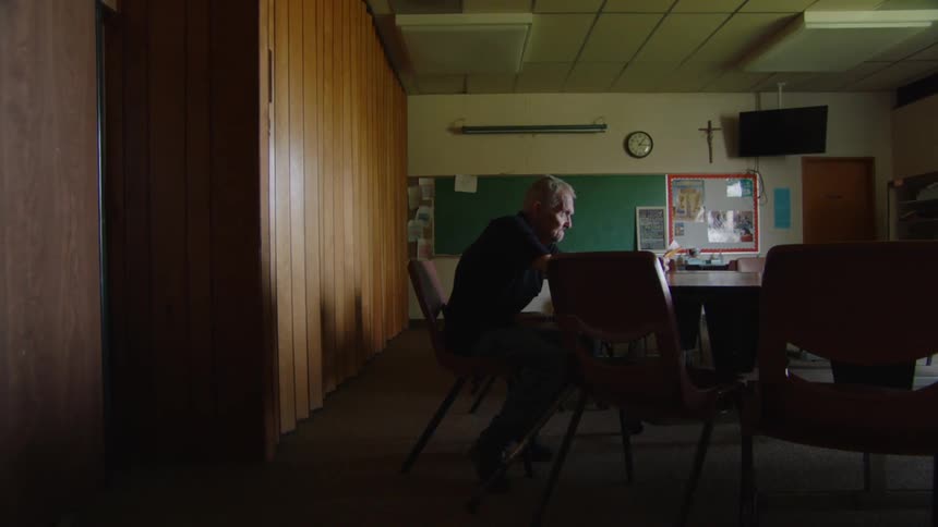 An elderly man sits alone in a dimly lit room reading a book