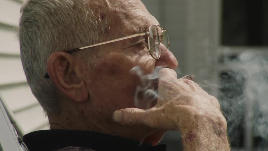 The video shows a closeup shot of an elderly mans hands resting on the armrests of what looks like a chair or walker he is wearing jeans and a cigarette is visible between his fingers the scene is outdoors on a sunny day